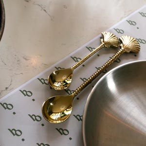 Pode incluir: Duas colheres douradas com um design de concha do mar no cabo. As colheres estão sobre um tapete de mesa branco e verde com uma tigela de prata no fundo.