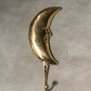 Puede incluir: Un gancho de pared de media luna dorado con una cara esculpida en la curva de la luna.