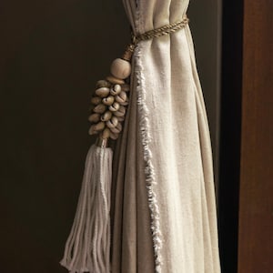 Può includere: Una tenda di lino bianco con un fermaglio a nappina realizzato con materiali naturali. La nappina è composta da piccole conchiglie e una perla di legno. La tenda ha un bordo sfrangiato.