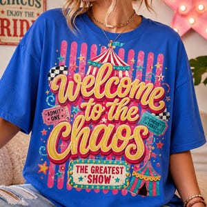 May include: A blue t-shirt with the words "Welcome to the Chaos" in yellow and pink, circus-themed graphics, and the text "The Greatest Show". The shirt features circus tent illustrations and star accents.
