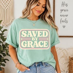 May include: A mint green t-shirt with the words "SAVED BY GRACE" in a retro-style design. The text is in a cream color with pink and light brown accents. The shirt is paired with blue denim jeans.