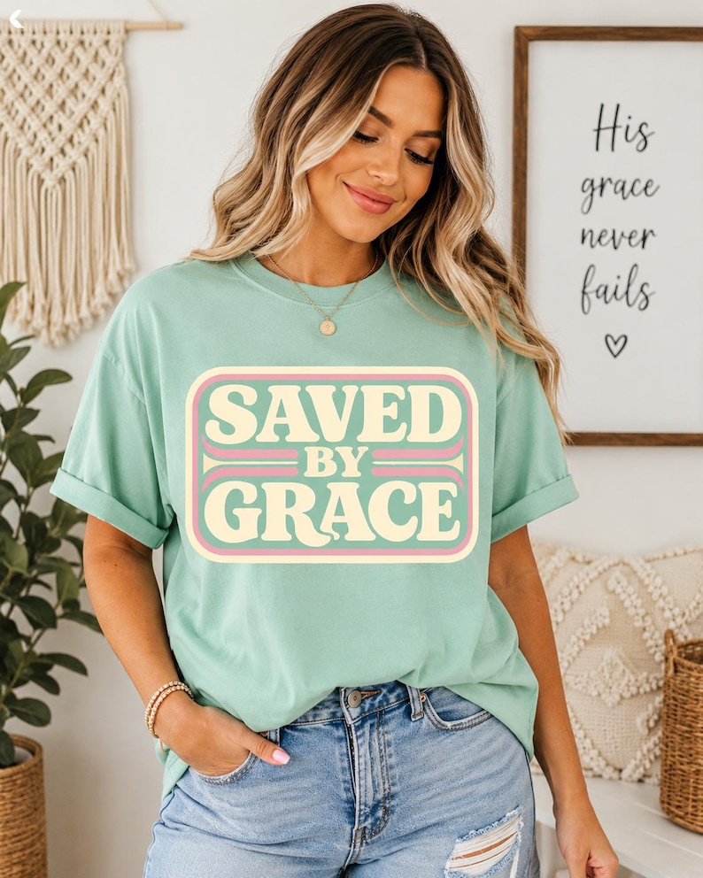 May include: A mint green t-shirt with the words "SAVED BY GRACE" in a retro-style design. The text is in a cream color with pink and light brown accents. The shirt is paired with blue denim jeans.