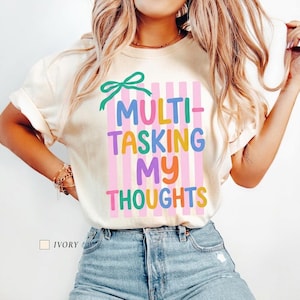 May include: An ivory short-sleeve t-shirt featuring a green bow graphic above vertical pink stripes and the multi-coloured text "MULTI-TASKING MY THOUGHTS" in a playful font. The shirt is paired with blue denim jeans, creating a casual look.