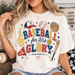 Puede incluir: Una camiseta color crema con un estampado gráfico que incluye el texto BASEBALL for His GLORY y 1 CORINTHIANS 10:31. El diseño presenta ilustraciones de una gorra de béisbol, una pelota, un guante, un bate, un megáfono y un dedo de espuma.