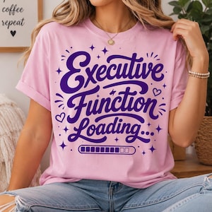 May include: A light pink short-sleeved t-shirt featuring dark purple text that reads Executive Function Loading... with a progress bar graphic, small heart icons, and star accents printed on the centre of the chest area.