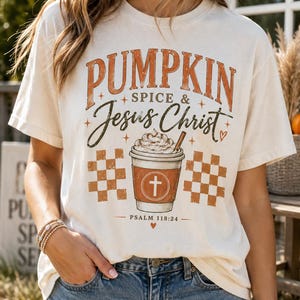 May include: An off-white short-sleeve t-shirt featuring a graphic print with the text PUMPKIN SPICE & JESUS CHRIST, a drawing of a coffee cup with whipped cream, two orange checkered squares, and the text PSALM 118:24 at the bottom.