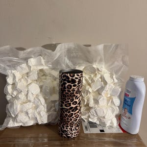 Puede incluir: Un vaso de color marrón y blanco con estampado de leopardo, una bolsa de cacahuetes de embalaje blancos y una botella blanca de talco para bebés con una etiqueta azul que dice "Johnson's Baby Powder".
