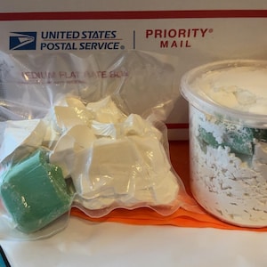 Puede incluir: Dos paquetes que contienen barras de jabón blancas y verdes. Los paquetes están etiquetados con "PRIORITY MAIL" y "UNITED STATES POSTAL SERVICE".