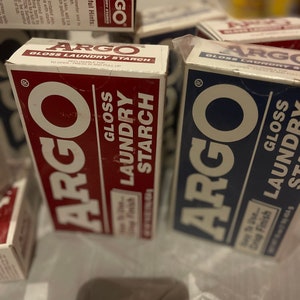 Puede incluir: Se ven varias cajas de almidón para ropa Argo Gloss, con embalajes rojos y azules. Las cajas tienen texto blanco y el logotipo de Argo. Las cajas están sobre una superficie blanca.