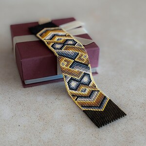 May include: A wide woven bead bracelet featuring a geometric pattern in shades of metallic gold, charcoal gray, and white. The bracelet has solid black beaded ends and is displayed resting on a square, dark maroon gift box with a light ribbon.