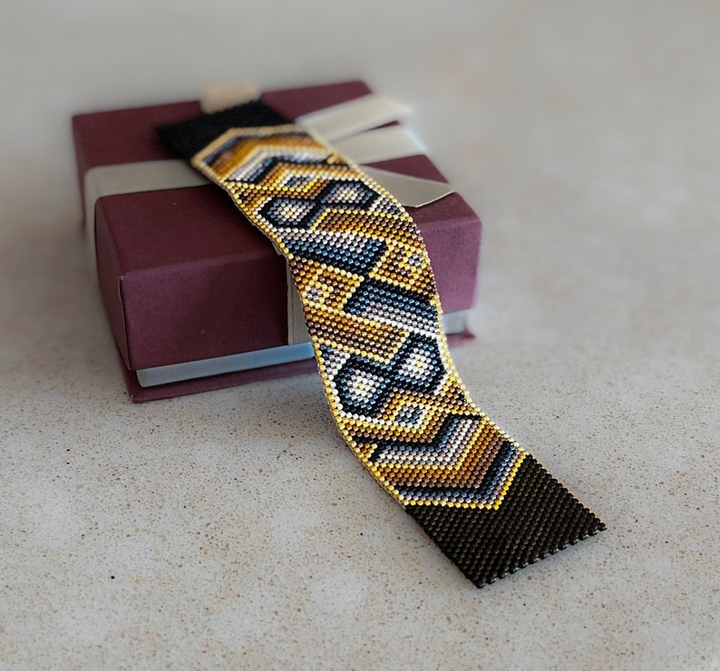 May include: A wide woven bead bracelet featuring a geometric pattern in shades of metallic gold, charcoal gray, and white. The bracelet has solid black beaded ends and is displayed resting on a square, dark maroon gift box with a light ribbon.