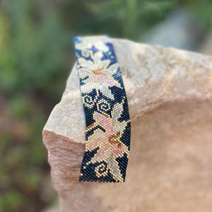 以下が含まれることがあります： 花柄のデザインのビーズブレスレット。ブレスレットは、ピンク、ゴールド、白の花柄のダークブルーの背景が特徴です。ブレスレットは小さくカラフルなビーズで作られており、石の表面に表示されています。
