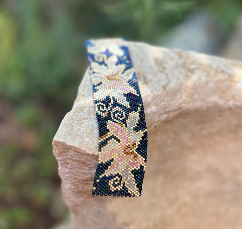 以下が含まれることがあります： 花柄のデザインのビーズブレスレット。ブレスレットは、ピンク、ゴールド、白の花柄のダークブルーの背景が特徴です。ブレスレットは小さくカラフルなビーズで作られており、石の表面に表示されています。