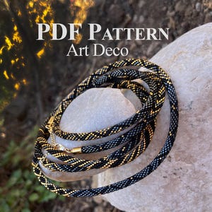 Könnte beinhalten: Eine schwarze und goldfarbene Perlenkette mit einem goldenen Verschluss, auf einem hellgrauen Stein aufgerollt. Die Halskette hat ein sich wiederholendes Muster aus schwarzen, goldenen und grauen Perlen. Der Text "PDF PATTERN Art Deco" ist oben links zu sehen.