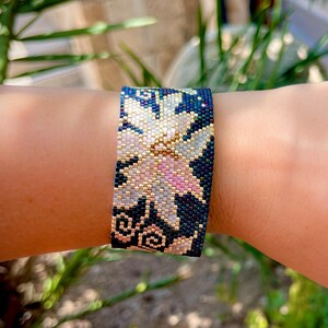 以下が含まれることがあります： 花柄のビーズブレスレット。白、ピンク、金色の花が濃い青色の背景に描かれています。精巧なビーズ細工がテクスチャード加工された表面を作り出し、ジュエリーの職人技を際立たせています。