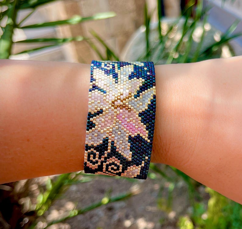以下が含まれることがあります： 花柄のビーズブレスレット。白、ピンク、金色の花が濃い青色の背景に描かれています。精巧なビーズ細工がテクスチャード加工された表面を作り出し、ジュエリーの職人技を際立たせています。