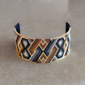 May include: A wide cuff bracelet crafted from small glass beads in a geometric pattern. The design features repeating diamond and chevron shapes in a color palette of metallic gold, dark navy blue, charcoal gray, and light silver, with a gold border.