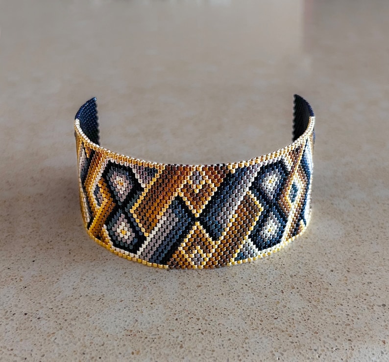 May include: A wide cuff bracelet crafted from small glass beads in a geometric pattern. The design features repeating diamond and chevron shapes in a color palette of metallic gold, dark navy blue, charcoal gray, and light silver, with a gold border.