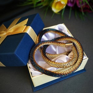 Op de afbeelding: Een gouden en zwarte kralenketting in een blauwe geschenkdoos met een gouden lint. De ketting is opgerold en heeft een metallic glans.