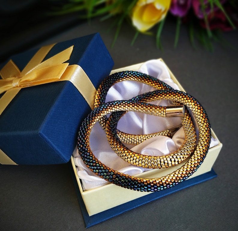 Op de afbeelding: Een gouden en zwarte kralenketting in een blauwe geschenkdoos met een gouden lint. De ketting is opgerold en heeft een metallic glans.