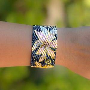 以下が含まれることがあります： 花柄のビーズブレスレット。ブレスレットは、濃い青色の背景に、黄色、ピンク、白のビーズのパターンが特徴です。ブレスレットの幅は約3.8cmです。