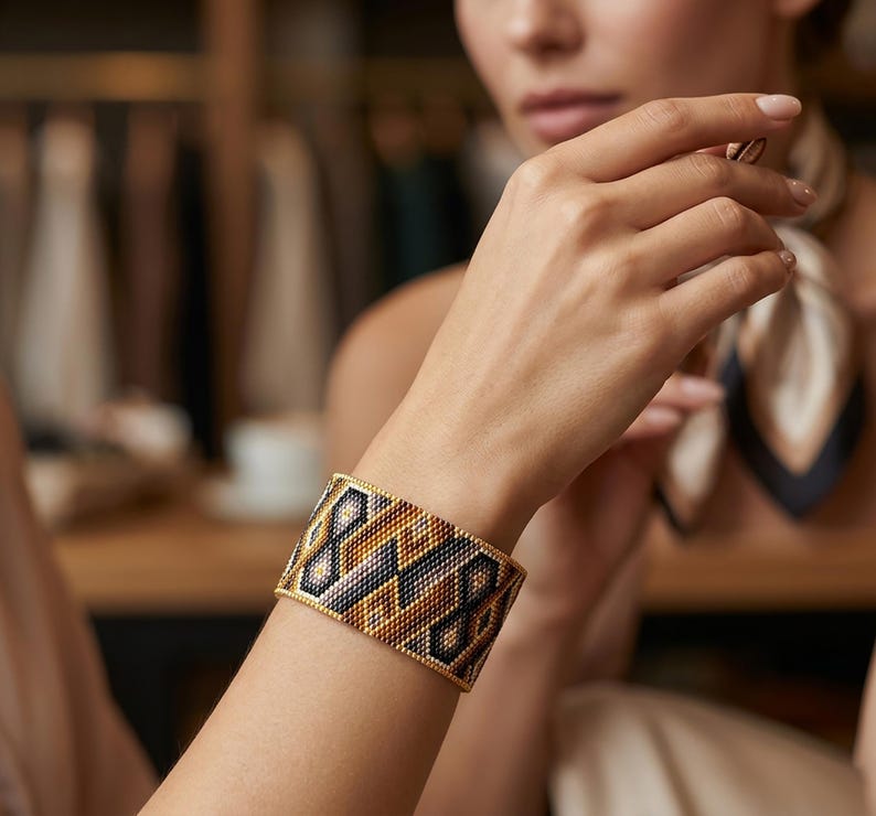 May include: A wide cuff bracelet crafted from small beads in a geometric pattern. The design features repeating diamond and chevron shapes in a color palette of metallic gold, deep black, and soft beige, creating a structured and textured appearance.