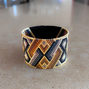 May include: A wide cuff bracelet crafted from tiny glass beads in a geometric pattern. The design features repeating diamond and triangular shapes in a color palette of metallic gold, bronze, charcoal gray, and black, with a gold beaded edge.