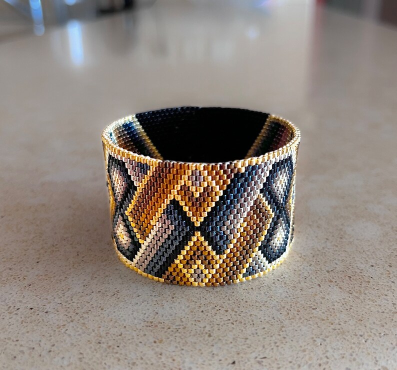 May include: A wide cuff bracelet crafted from tiny glass beads in a geometric pattern. The design features repeating diamond and triangular shapes in a color palette of metallic gold, bronze, charcoal gray, and black, with a gold beaded edge.