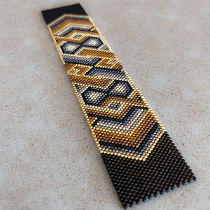 May include: A rectangular beaded bracelet featuring a geometric pattern in metallic gold, silver, and matte black glass beads. The design consists of repeating chevron and diamond shapes arranged in a vertical orientation with a flat, woven texture.