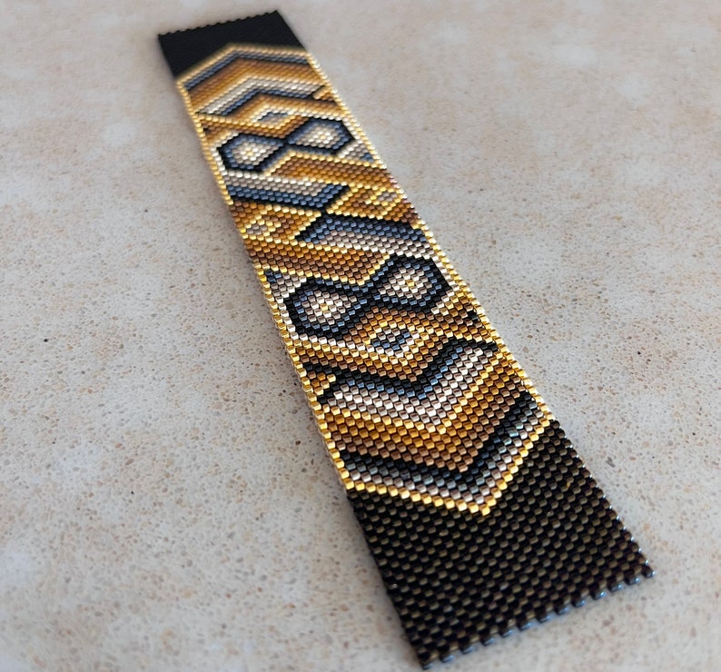 May include: A rectangular beaded bracelet featuring a geometric pattern in metallic gold, silver, and matte black glass beads. The design consists of repeating chevron and diamond shapes arranged in a vertical orientation with a flat, woven texture.