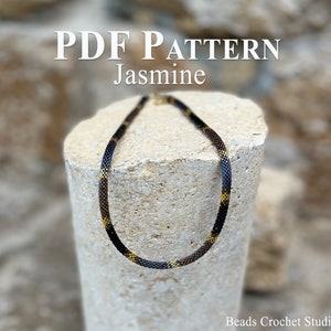 May include: PDF pattern for a beaded necklace called "Jasmine". The necklace is made with black and gold beads and is shown on a stone surface.