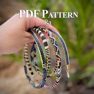 Könnte beinhalten: Eine lange, flexible Perlenketten-Schnur mit einem geometrischen Muster aus schwarzen, goldenen, weißen und roten Saatperlen. Die Kette hat einen goldfarbenen Metallverschluss an einem Ende. Der Text PDF PATTERN Ra ist oben im Bild zu sehen.