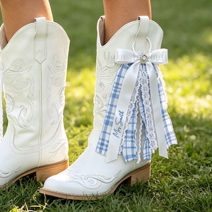Puede incluir: Un par de botas vaqueras de cuero blanco con bordados tonales. La bota derecha está decorada con un conjunto de cintas que incluye tela de cuadros azules y blancos, encaje blanco y una cinta blanca con el texto Mrs Smith impreso.
