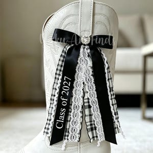 May include: A white western boot featuring decorative stitching and a layered ribbon accessory. The accessory includes black satin, black and white gingham, and white lace ribbons, with a rhinestone center and text reading Class of 2027.
