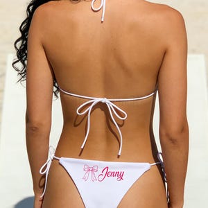 Può includere: Bikini bianco con lacci. Il top e il fondo del bikini sono bianchi. Il fondo ha una grafica a fiocco rosa e il nome "Jenny" stampato in rosa.