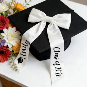 Può includere: Un cappello di laurea nero con un fiocco di raso bianco e nastri. Il nastro a sinistra dice "Emily" con un'icona del cappello di laurea, e il nastro a destra dice "Class of 2020" in corsivo nero. Un bouquet di fiori è sullo sfondo.