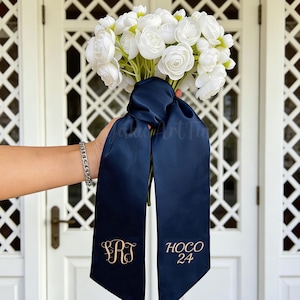 Könnte beinhalten: Ein Strauß weißer Blumen, gebunden mit einem marineblauen Satinband. Das Band hat goldgestickte Initialen und den Text "HOCO 24". Die Blumen werden vor einer weißen Tür gehalten.