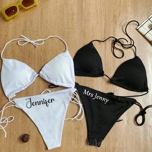 May include: Two bikinis, one white and one black, with matching bottoms. The white bikini bottom says "Jennifer" and the black bikini bottom says "Mrs Jenny". Yellow sunglasses are in the top left corner.