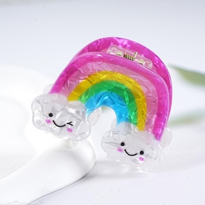 Op de afbeelding: Een regenboogkleurige haarspeld met een roze basis en heldere wolkaccenten. De wolken hebben glimlachende gezichten.
