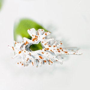 May include: White metal hair clip with a splatter paint design in orange. The clip is shaped like a seahorse.