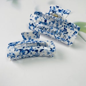K&ouml;nnte beinhalten: Zwei blaue und wei&szlig;e gemusterte Haarklammern. Die Klammern sind aus Kunststoff und haben ein Klauen-Design.