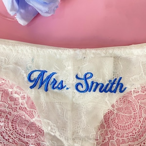 Könnte beinhalten: Weiße Spitzenunterwäsche mit dem in blauer Schrift gestickten Namen "Mrs. Smith". Die Unterwäsche hat ein zartes florales Spitzenmuster und ist vor einem rosa Hintergrund platziert. Oben links ist eine hellblaue Blume zu sehen.