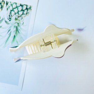 May include: A white plastic hair claw with a clear plastic spring mechanism. The claw has a smooth, rounded shape and a textured grip.