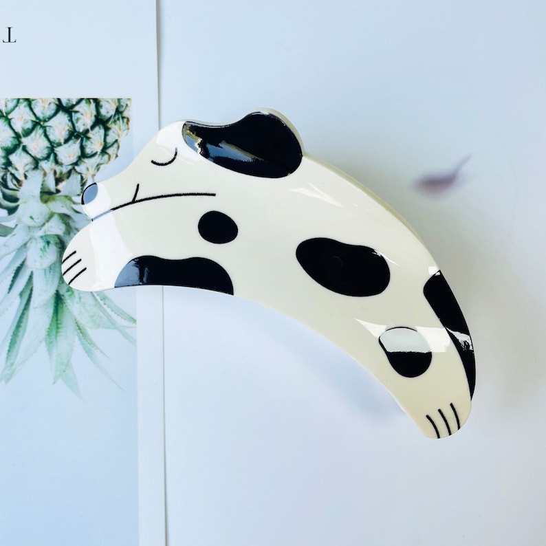 May include: A white ceramic hair clip shaped like a sleeping dalmatian dog with black spots. The clip is curved and has a glossy finish.