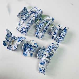 K&ouml;nnte beinhalten: Satz mit sechs blauen und wei&szlig;en Haarklammern mit Muster. Die Klammern sind aus Kunststoff und haben verschiedene Formen, darunter Schmetterling, Rechteck und Krabbe.