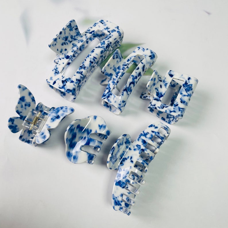 K&ouml;nnte beinhalten: Satz mit sechs blauen und wei&szlig;en Haarklammern mit Muster. Die Klammern sind aus Kunststoff und haben verschiedene Formen, darunter Schmetterling, Rechteck und Krabbe.