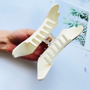 May include: A white plastic hair claw with a clear plastic spring mechanism. The claw is designed to hold hair in place.