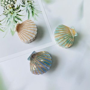Puede incluir: Tres pinzas para el pelo decorativas con forma de conchas. Las conchas son blancas, azules y azul claro con detalles dorados.