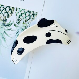 May include: A white and black spotted dog-shaped hair clip. The clip is curved and has a black outline. The dog has a black nose and eyes.