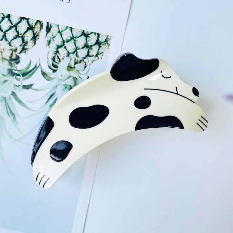 May include: A white and black spotted dog-shaped hair clip. The clip is curved and has a black outline. The dog has a black nose and eyes.
